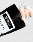 Pink Kursiv bath salts pouring onto a white surface from a black bag with label.