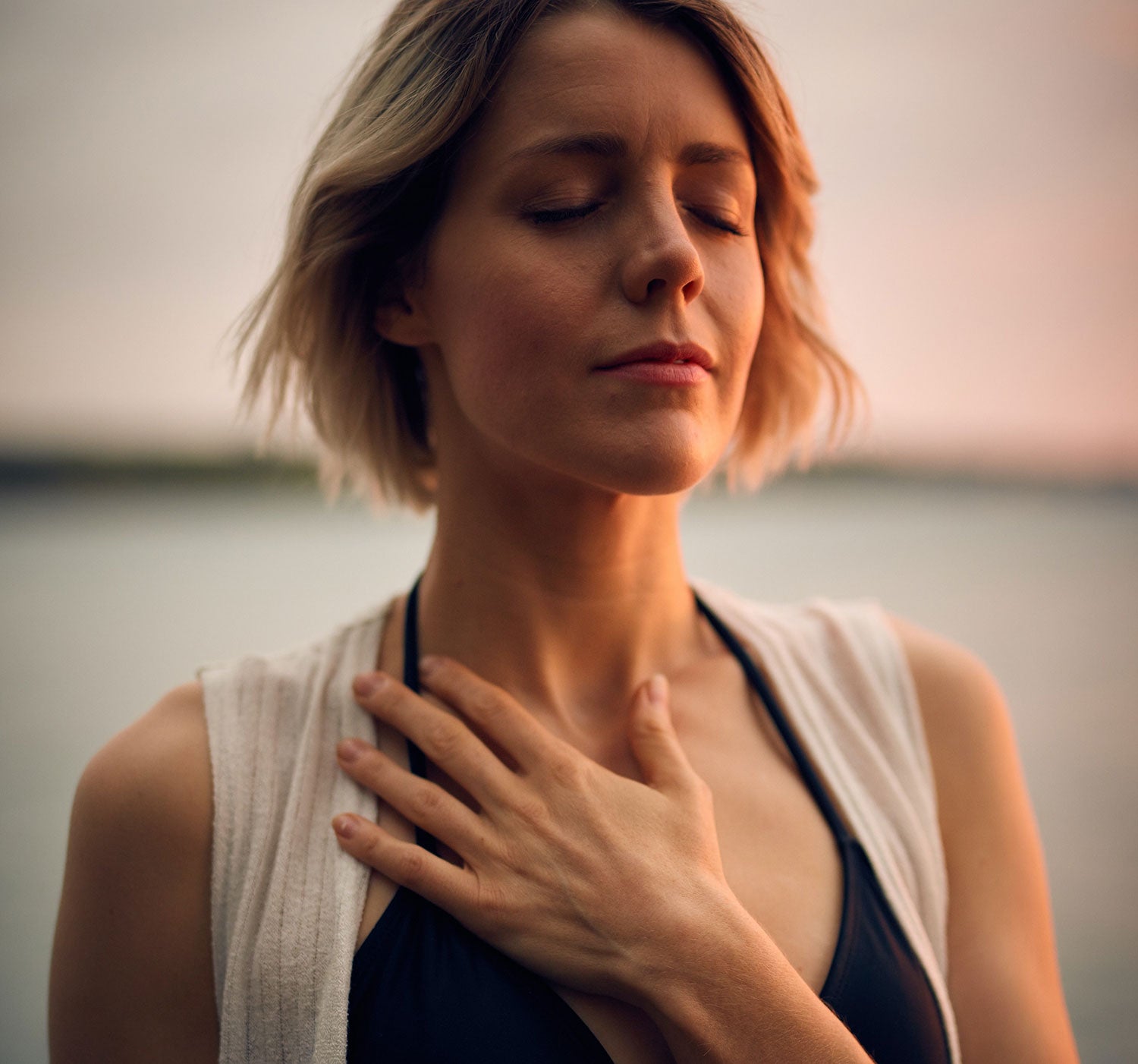 A blond woman in a black bikini top and white vest closes her eyes and places her hand on her chest. Image by Darius Bashar