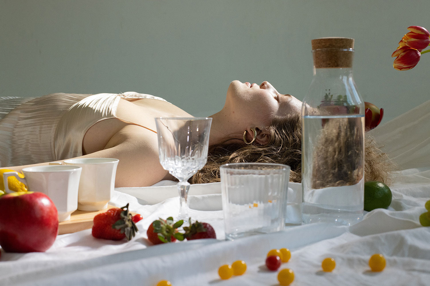 A peaceful young woman in a bra top lays with her eyes closed near assorted glassware and fresh fruit on a blanket. Image by Jill Burrow.