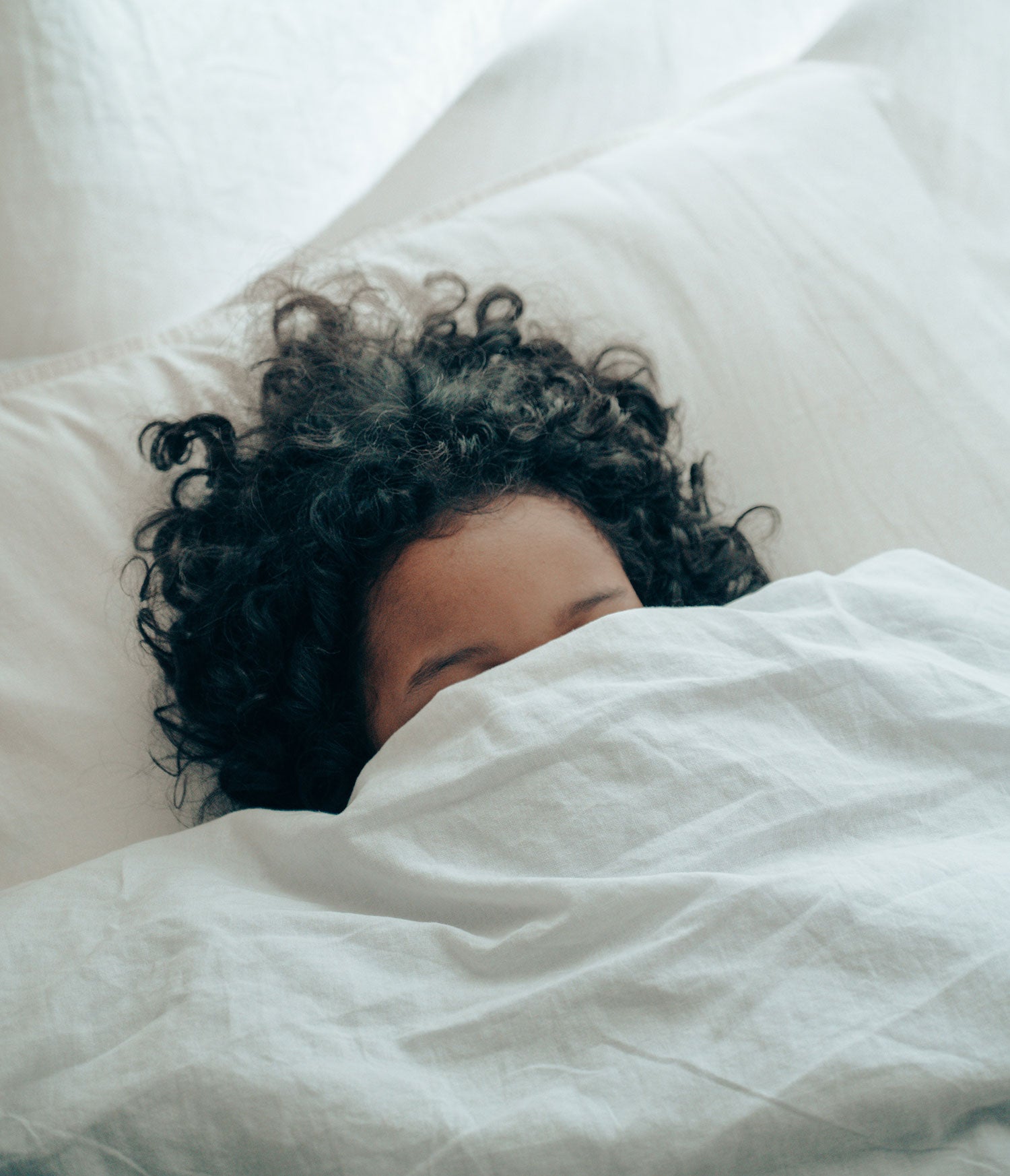 An unrecognizable person with black curly hair sleeps under a white blanket on a white pillow. Image by Ketut Subiyanto.