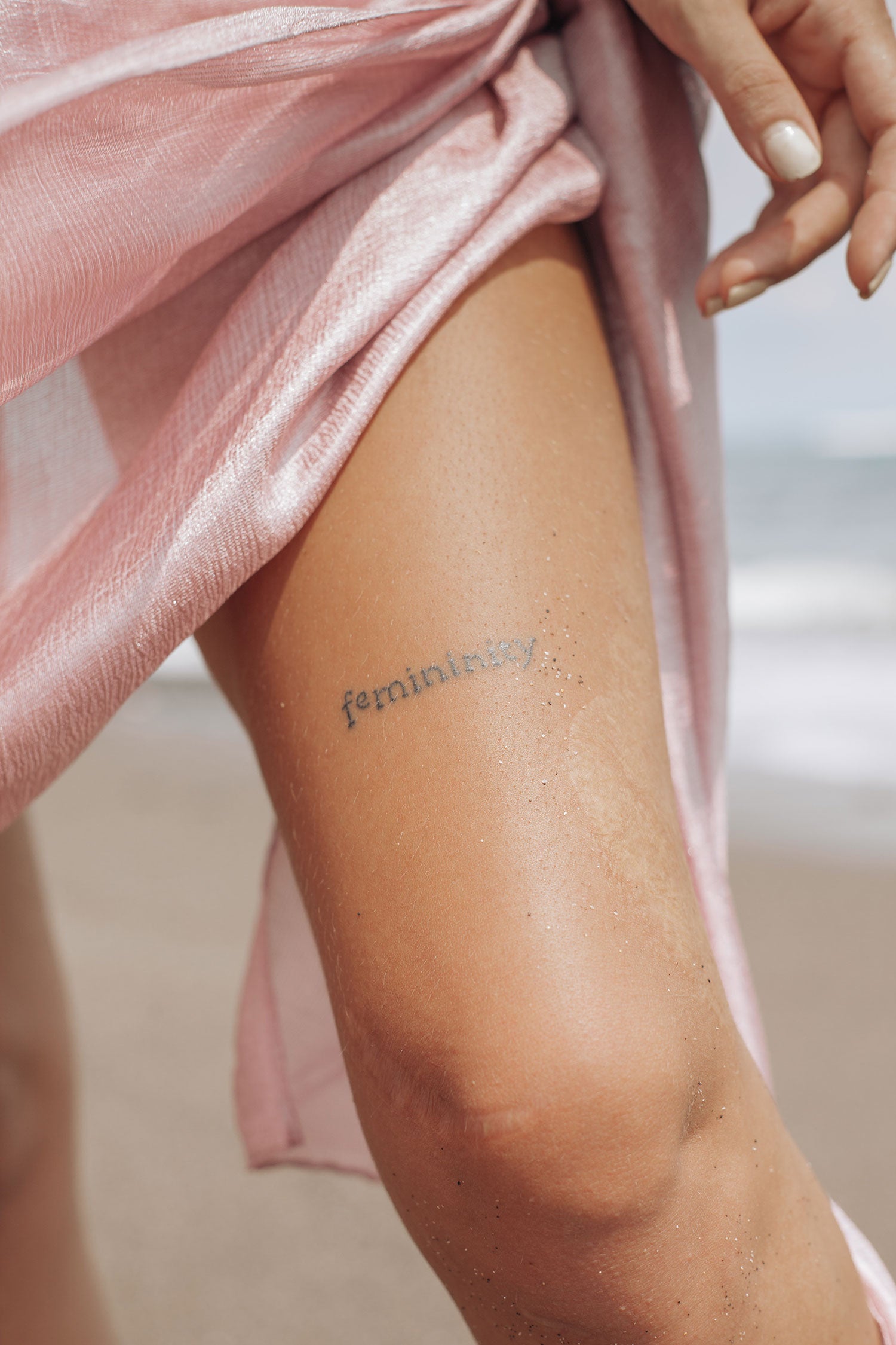 A woman with the word femininity tattooed on her thigh walks on the beach in a pink coverup. Image by Ksu&Eli.