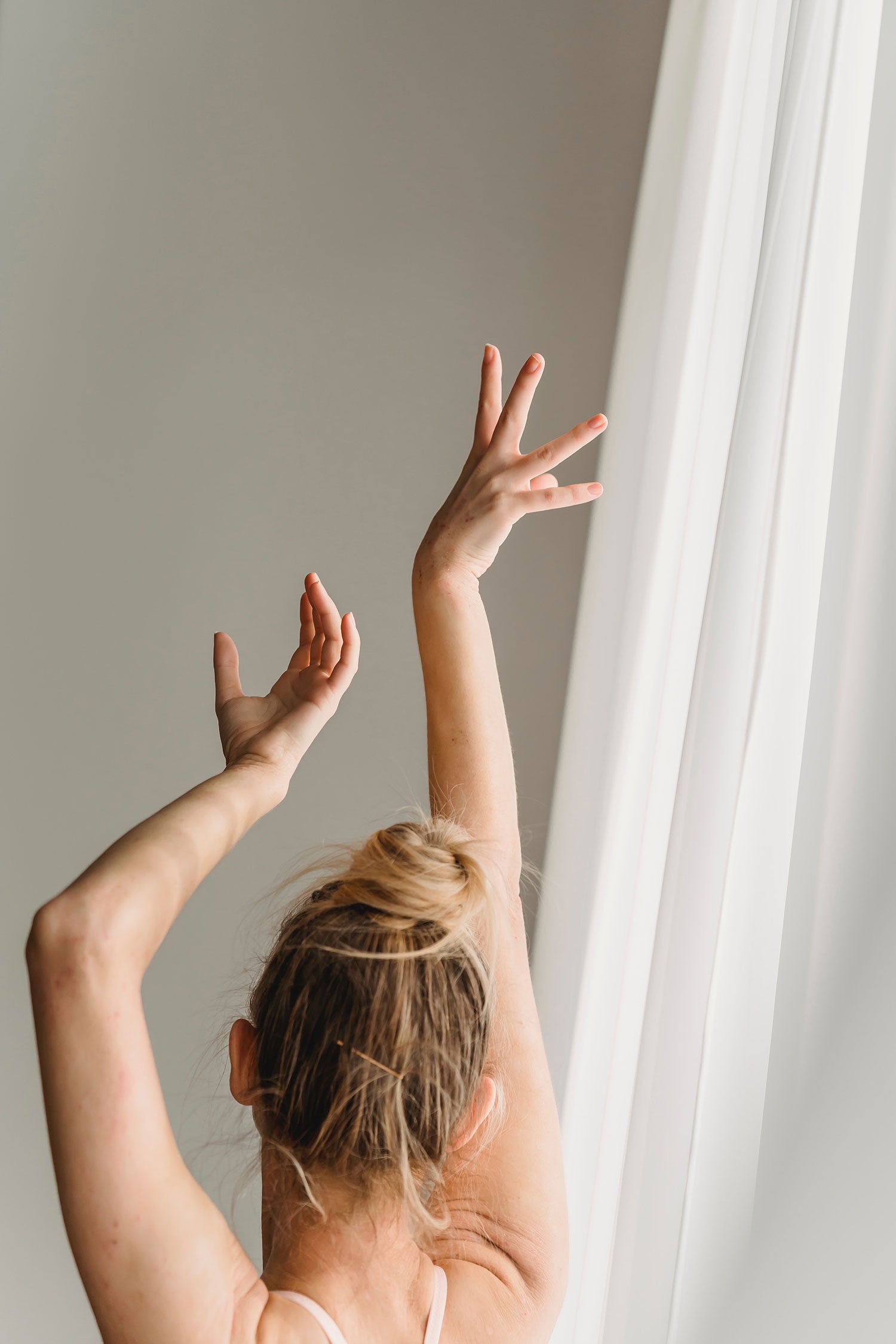 A white woman with her blond hair in a bun holds her arms above her head dramatically next to a gauzy white curtain. Image by Marta Wave.