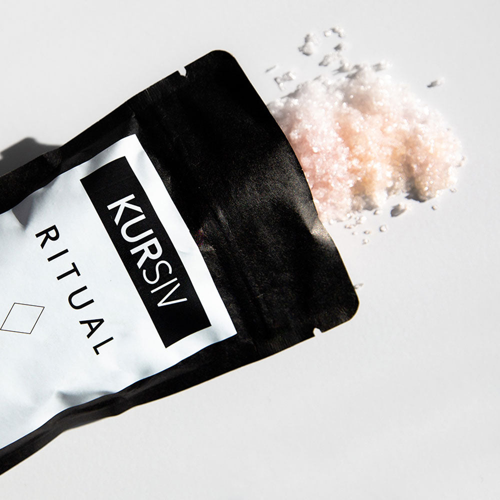 Pink Kursiv bath salts pouring onto a white surface from a black bag with label.